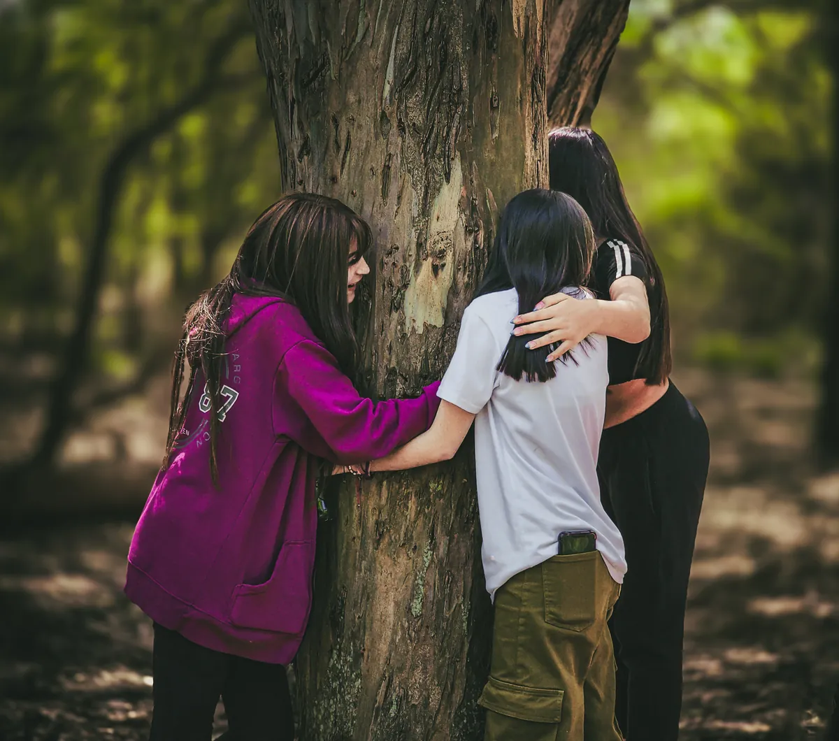 Tres chicas de espaldas abrazan el tronco de un árbol